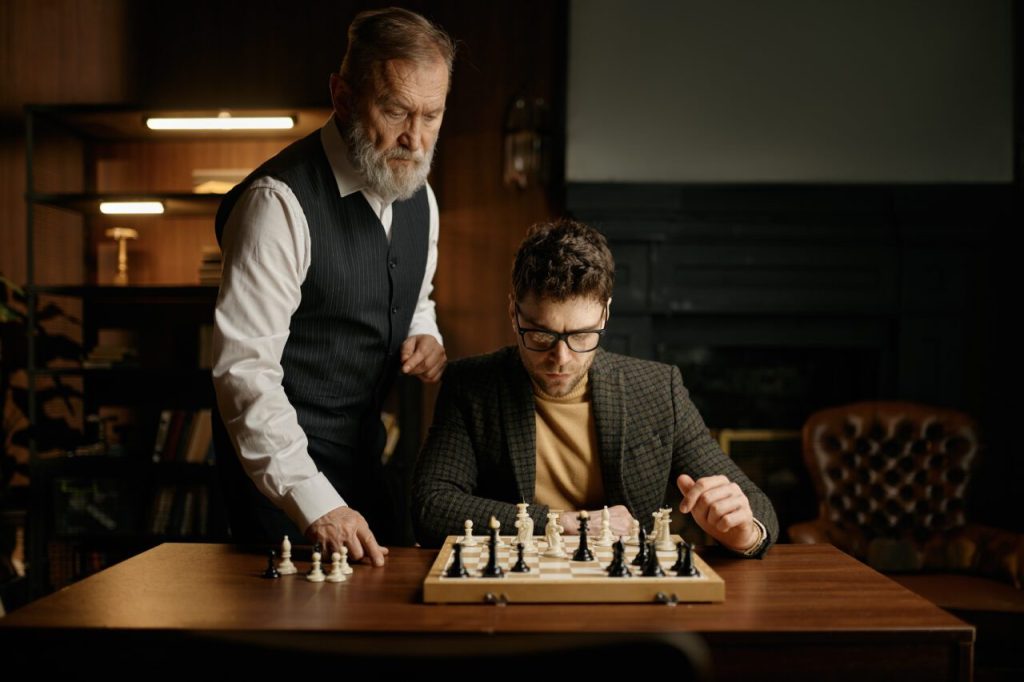 Senior man teaching young guy playing chess explaining rules of game 1 e1698221466620.jpg
