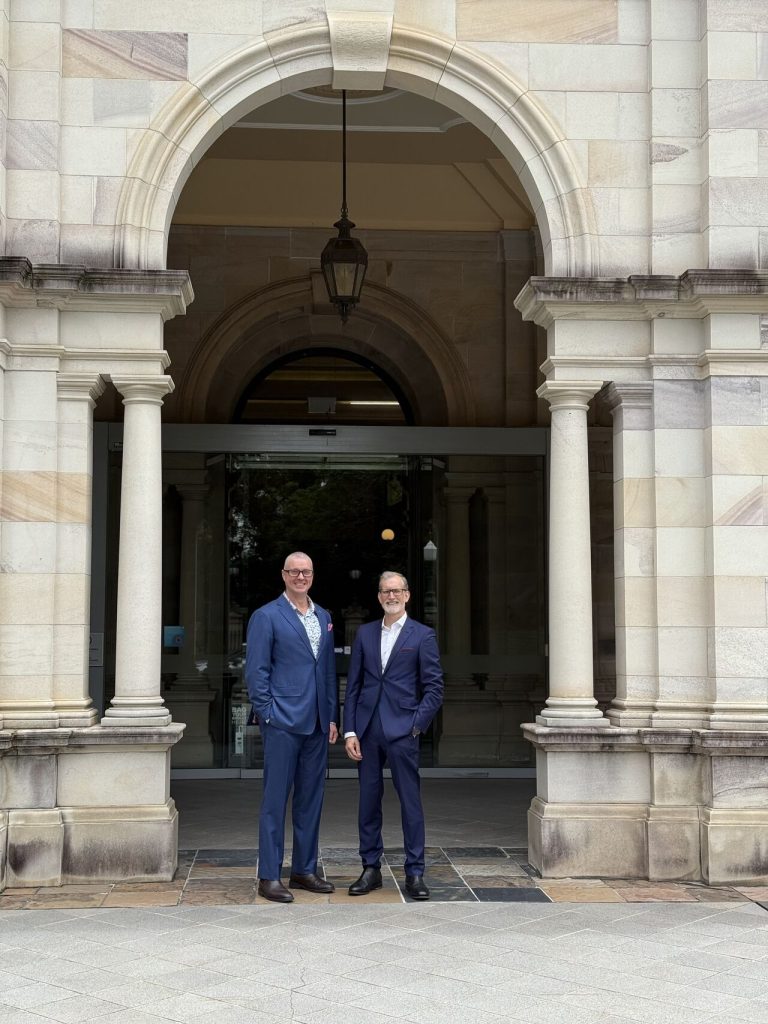 Two men standing in archway of an old building wearing navy blue suits.